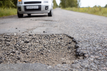 Country road in terrible condition. Modern grey vehicle approaching huge pothole