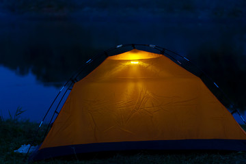 An orange tent at night stands on the shore and glows inside.