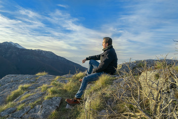 Yong man enjoying scenic view and sunset in mountains sitting on rocky floor. Success and freedom concept. Copy space.