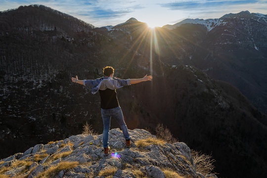 Happy motivated and inspired man with spread hands stands on the edge of rocky cliff watching sunset in the mountains. Motivation and inspiration concept. Freedom concept. - Powered by Adobe
