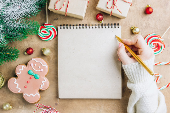 Notebook in the center of cute Christmas decoration: gift box, christmas balls, gingerbread man, fir tree branch, cande cane. Female hand writing in notebook. Flat lay. Top view