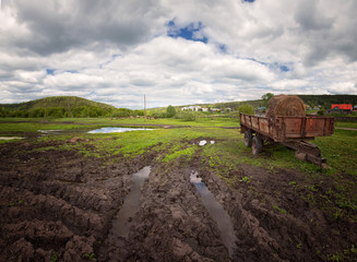 hay season, summer landscape old iron trailer with a roll of hay on a dirty village road, white clouds in the blue sky,
