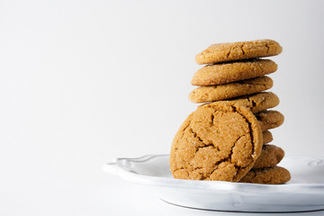 A ginger cookie leans against a stack of ginger cookies on a white plate and a white background with copy space