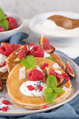 Pancakes with raspberries, figs, yogurt, coconut zest, honey and mint leaves on a plate.
