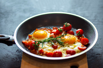 Fried scrambled eggs in a pan on a black background with cherry tomatoes, cheese and rosemary. Fried egg with a liquid yolk is crushed by herbs. Eggs in a teflon pan. Quick breakfast for every day