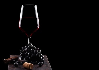 Glass of red wine with dark grapes and vintage corkscrew opener and cork on wooden board on black...