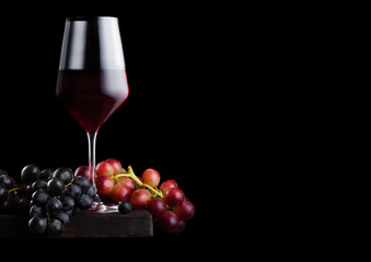 Elegant glass of red wine with dark and red grapes on wooden board on black background. Space for text