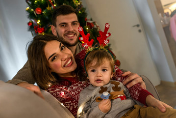 Joyful family enjoying on Christmas Eve at home. Happy young parents with their baby boy enjoy Christmas time while relaxing on the sofa. Family prepare for Christmas