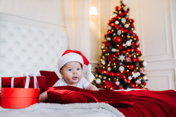 Fototapeta na wymiar Baby in costume of Santa Claus with gift box lie on the bed in Christmas living room.