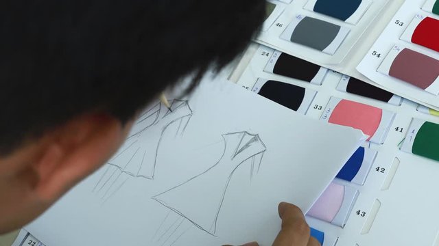 Close up scene video of Asian male fashion designer sketching his new collection during choosing material from fabric catalog in his studio.