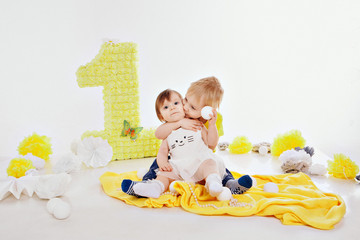 Birthday celebration: girl and boy sitting on the floor among the decoration: numbers 1, artificial flowers and white balls