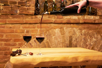Hand of a sommelier pouring red wine into long-stemmed wineglasses. Two wineglasses on wooden table in the old vintage wine cellar from red brick