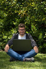 Young man sitting on the grass of a park and looking at the screen of his laptop while he is smiling. Natural Environment