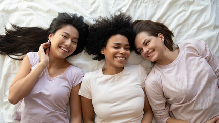 Three happy funny multiethnic ladies friends laughing lying on bed