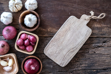 Top view of herbal vegetable ingredients, garlic, red onion, and empty chopping board on old wooden table, cooking preparation concept