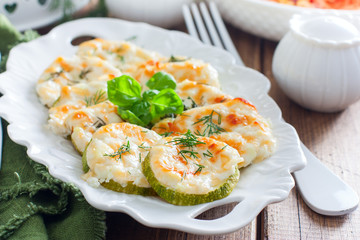 Baked zucchini with cheese slices on a white dish on a wooden table, horizontal