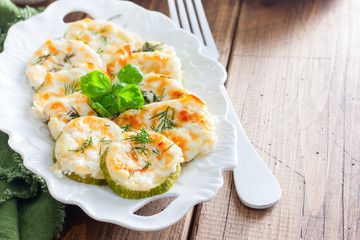 Baked zucchini with cheese slices on a white dish on a wooden table, horizontal, copy space