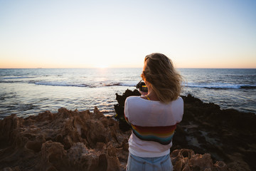 Young woman with blonde hair blowing in the wind, standing on rocks at Sunset taking photos of the...