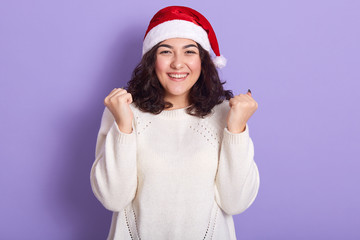 Beautiful female model wearing red santa claus and white sweater, raising her fists, smiling sincerely, having curly black hair, standing isolated over lilac background in studio, waiting for holiday.