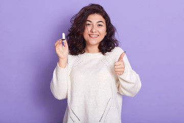 Close up portrait of female holds tampon over lilac background, having happy facial expression, shows ok sign, being glad with her choosing, has period, lady wears white shirt, wearing stylish outfit.