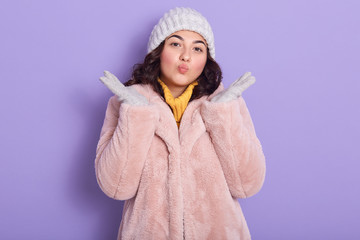 Eco friendly lover of animals raising hands, putting lips together, wearing pink fauxfur coat, yellow sweater, gloves and hat, standing isolated over lilac background in studio. Lifestyle concept.