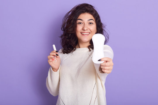 Attractive Caucasian woman holds cotton tampon and sanitary napkin, female prefer to use pads, has happy look, monthly menstrual cycle, lady wearing white sweater. Protective care for women health.