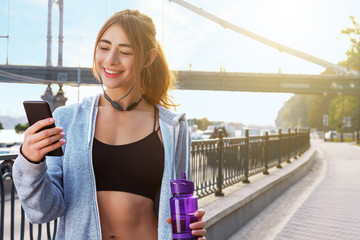 Young smiling woman is listening music and drinking water after morning exercises at street of city. Girl in wireless headphones is resting after jogging. Sporty and healthy lifestyle concept.