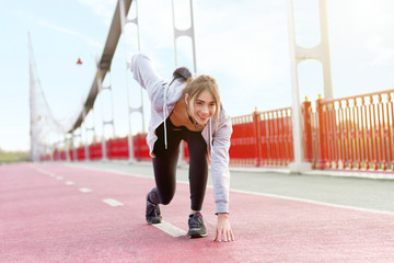 Young smiling woman is standing at starting position on urban bridge. Girl in sportswear is doing exercises, preparing for marathon running, jogging. Sporty and healthy lifestyle concept.