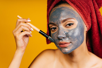 the girl applies a charcoal mask with a cosmetic brush on her face and carefully looks at the camera, as in a mirror