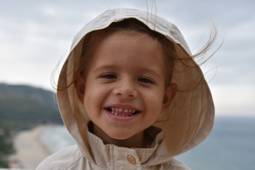 A little smiling girl is wearing a raincoat jacket with hood against the stormy sky. Childhood concepts. Environmental concepts. Climate concepts.