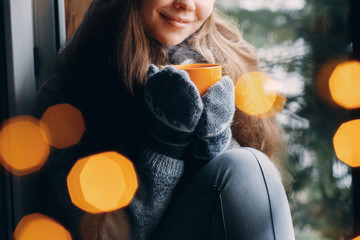 Woman's hand holding a cup of coffee or cocoain in gloves sitting home by the window. Blurred winter snow tree background. Morning, coziness, winter and people concept
