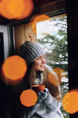 Beautiful  woman holding and drinking a cup of coffee or cocoain in gloves sitting home by the window. Blurred winter snow tree background. Morning, coziness, winter and people concept