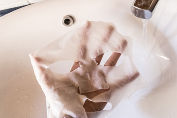 Woman hand holding a white sheet asian mask over a sink with the running water, skin care concept, washing sheet mask - Image