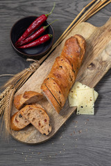 A fresh sliced rye-wheat flour baguette with bran on a wooden board and three slices of cheese and a bowl with three pepperoni pods next to it. Top view, with copy space