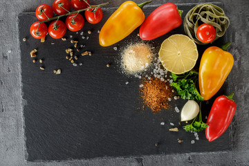Multicolored peppers, branch of fresh cherry tomatoes, pasta spinach fettuccine, parsley, slice of lemon and spices on a stone board. Top view, with copy space