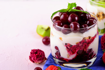 dessert trifle with cherries, yogurt and granola in glass on blue cloth napkin on light pink background
