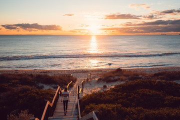 Young woman walking down the beach staircase and path to the sea with the orange sunset over the beautiful ocean in Perth, Western Australia.   - obrazy, fototapety, plakaty