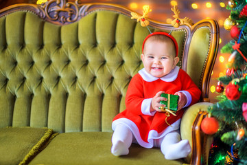 Cute little girl in a red dress is sitting on the sofa with a gift box in her hands and smiling. Christmas and New Year