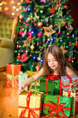 A cute girl in a white dress is sitting on the floor with a large pile of gifts. Against the background of a festive tree. Christmas and New Year