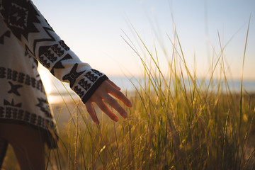 Young woman wearing a jersey with her hand exploring the sand dune grass on the beach with the sunset in the background over the ocean, Cottesloe, Perth, Western Australia.
