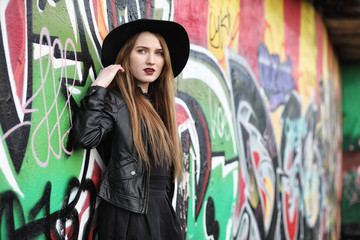 Obraz premium Young beautiful girl in a hat and with a dark make-up outside. Girl in the Gothic style on the street. A girl walks down the city street in a leather waistcoat with a phone.