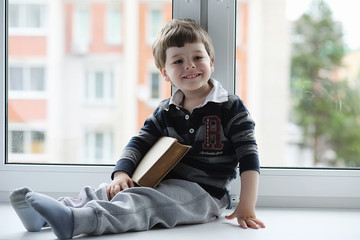 The little boy is reading a book. The child sits at the window and prepares for lessons. A boy with a book in his hands is sitting on the windowsill.