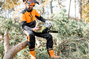 Obraz premium Portrait of a professional lumberman in protective workwear sitting with mobile phone on the felled tree, resting after the hard work in the forest