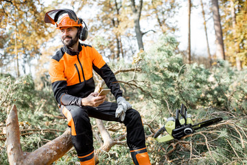 Portrait of a professional lumberman in protective workwear sitting with mobile phone on the felled...