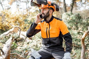 Professional lumberman in protective workwear talking on phone, while sitting on the felled tree, resting after the hard work in the forest