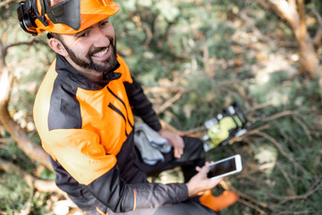 Portrait of a professional lumberman in protective workwear sitting with mobile phone on the felled tree, resting after the hard work in the forest