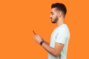 Side view of serious calm brunette man with beard in white t-shirt using cellphone, texting...