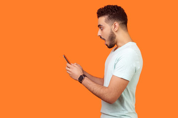 Side view of amazed brunette man with beard in white t-shirt looking shocked at mobile phone, pleasantly surprised by good gadget, application. empty copy space for advertisement, indoor studio shot