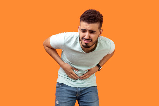 Stomachache. Portrait of sick young man with beard in casual white t-shirt grimacing and suffering from pain in belly, severe abdominal distress. indoor studio shot isolated on orange background