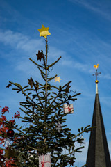 festlich dekorierter Weihnachtsbaum vor einem d&ouml;rflichen Kirchturm, blauer Himmer